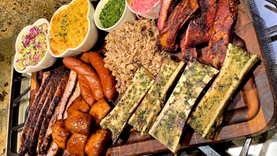 A wooden board filled with homemade BBQ meats, smoked sausages, ribs, brisket, pulled pork, roasted bone marrow, and sides like coleslaw, pickled onions, and green sauce.