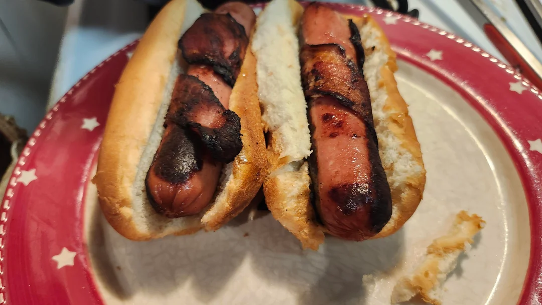 Two grilled hot dogs wrapped in crispy, charred bacon in buns are served on a red plate with white stars along the rim. A piece of bun sits nearby on the plate.