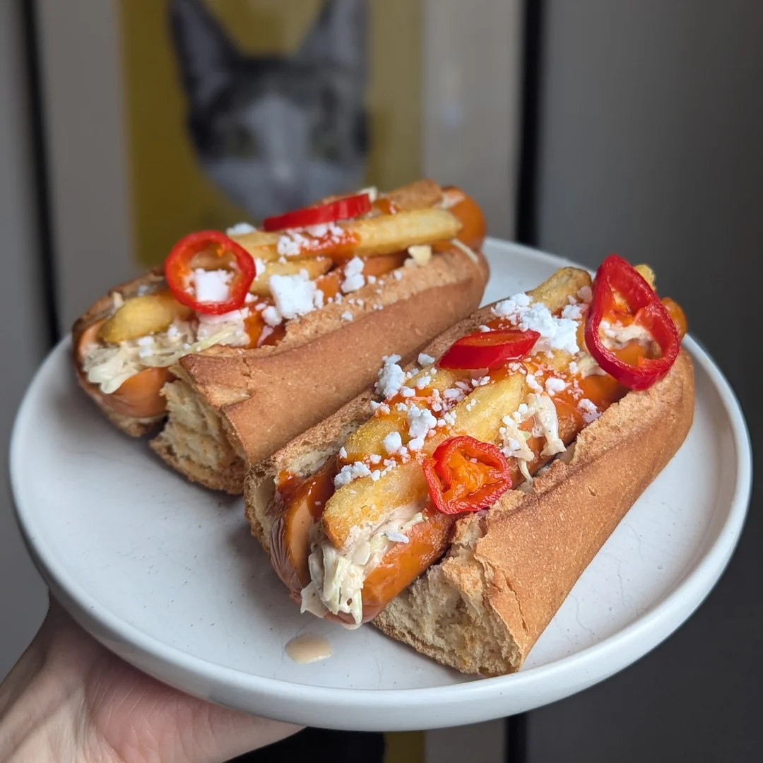 Two loaded hot dogs on a white plate, topped with fries, sliced red peppers, crumbled cheese, and sauce; a framed cat portrait is blurred in the background.