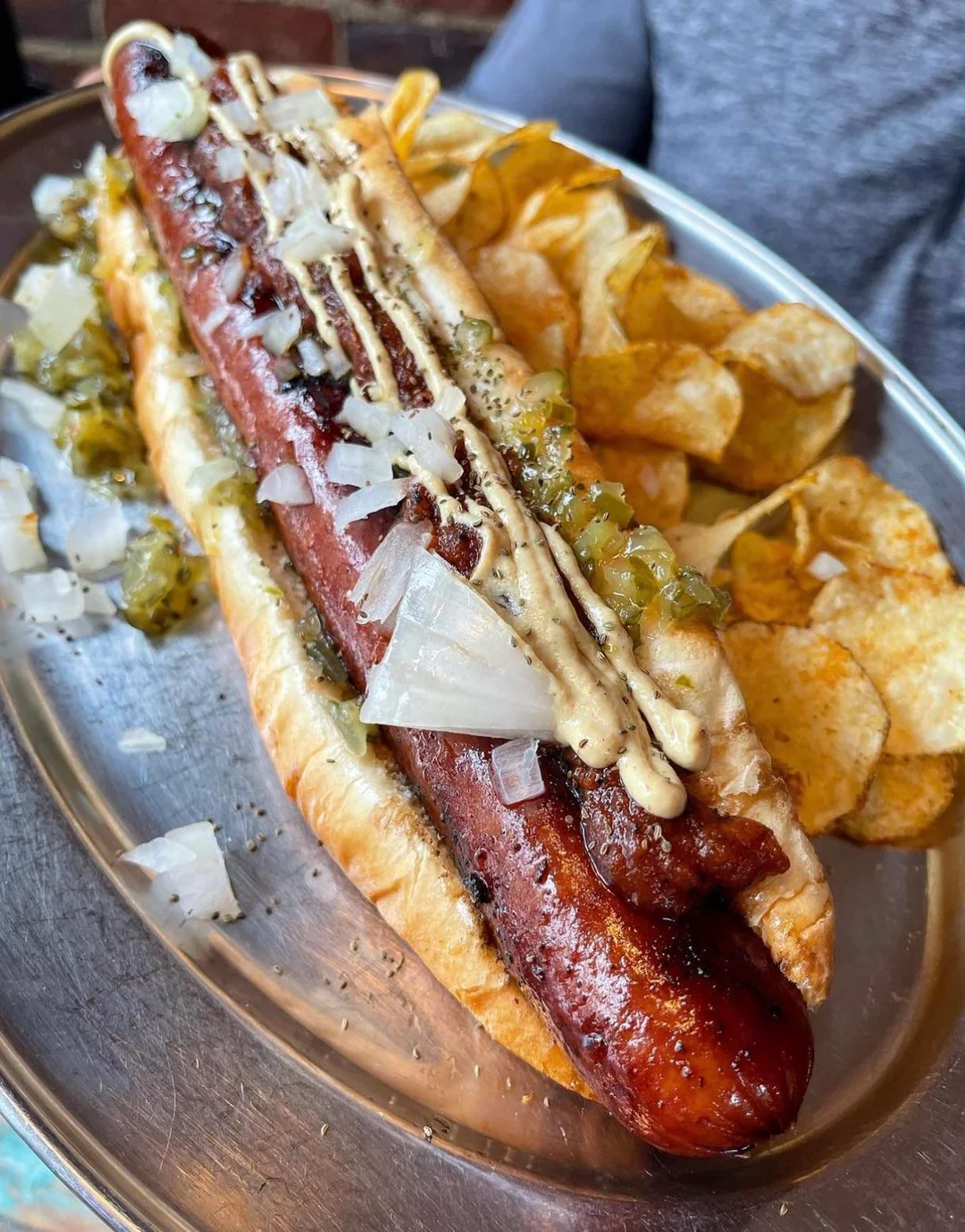 A grilled hot dog in a bun topped with chopped onions, relish, and drizzled mustard, served on a metal plate with a side of potato chips.