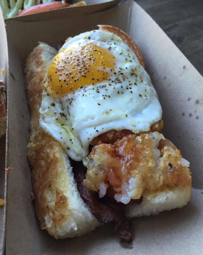 A sandwich in a cardboard tray topped with crispy bacon, a fried sunny-side-up egg, and a piece of deep-fried food on a toasted bun.