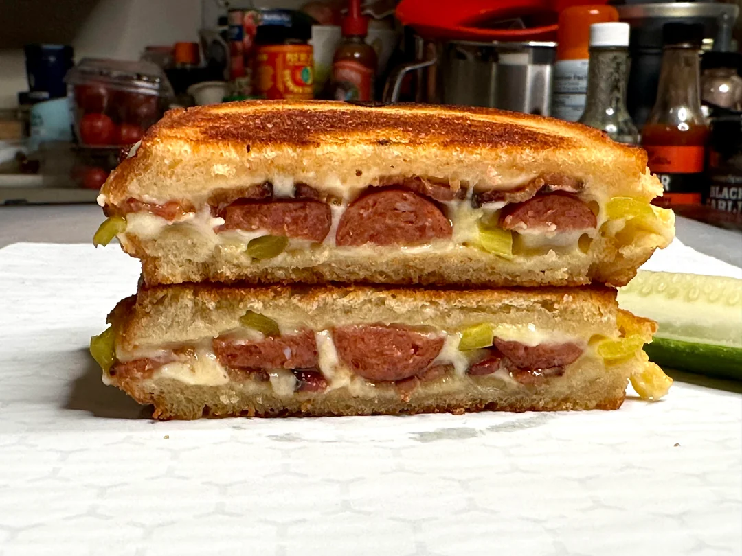A grilled cheese sandwich with sausage and pickles, cut in half and stacked, showing melted cheese, sausage slices, and pickles inside. A pickle spear is on the side, with kitchen items blurred in the background.