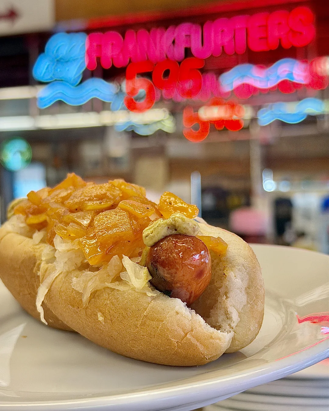 A hot dog with sauerkraut, onions, and mustard on a plate with a neon "FRANKFURTERS 5.95" sign blurred in the background.