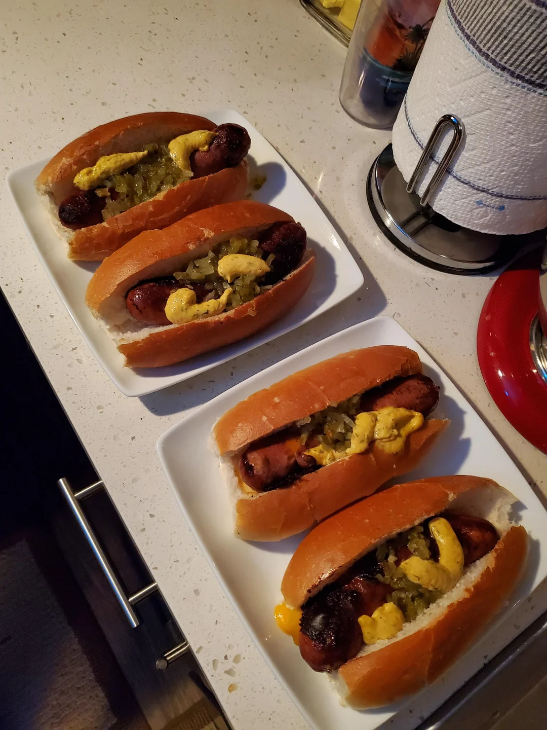 Four hot dogs with mustard, relish, and onions sit on white plates on a kitchen counter next to paper towels and appliances.