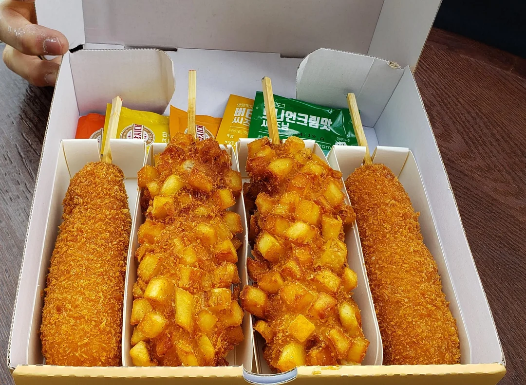 A box contains four Korean corn dogs on sticks, two with a diced potato coating and two with a classic golden breaded coating. Packets of mustard and sauce are included in the box.