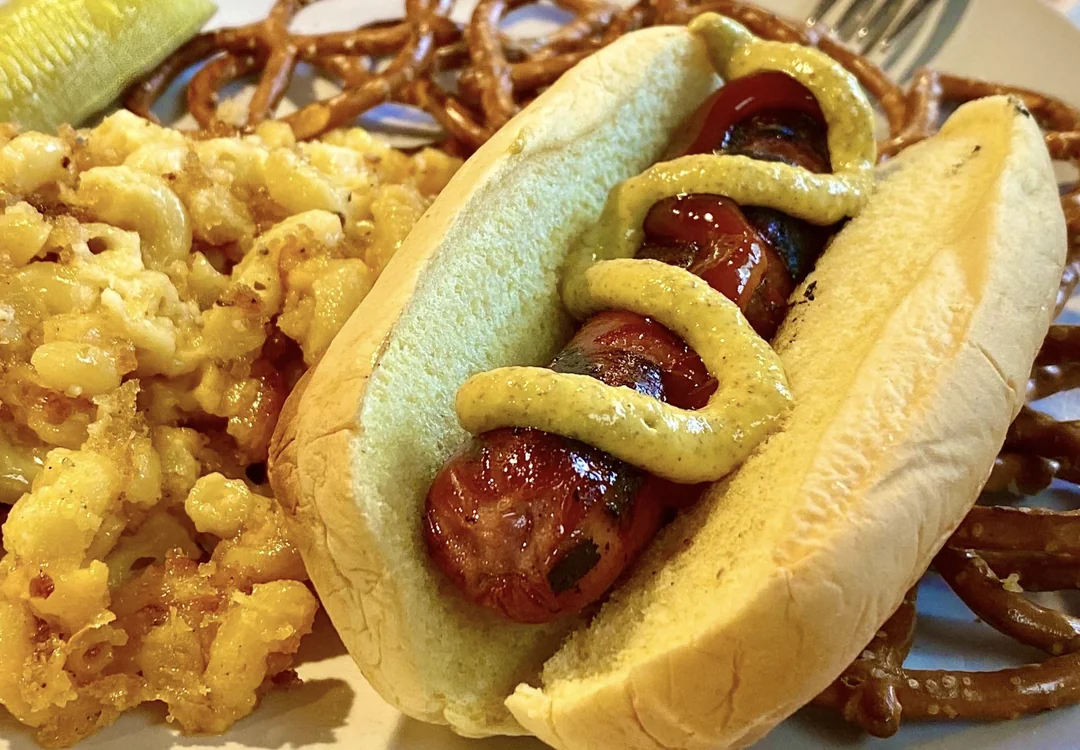 A grilled hot dog with mustard in a bun, served with macaroni and cheese, pretzels, and a pickle on a plate.