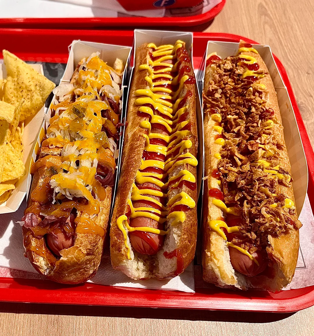 Three loaded hot dogs on a red tray: one with sauerkraut, cheese, and onions; one with ketchup and mustard zigzags; and one topped with crispy fried onions. Tortilla chips are visible on the side.
