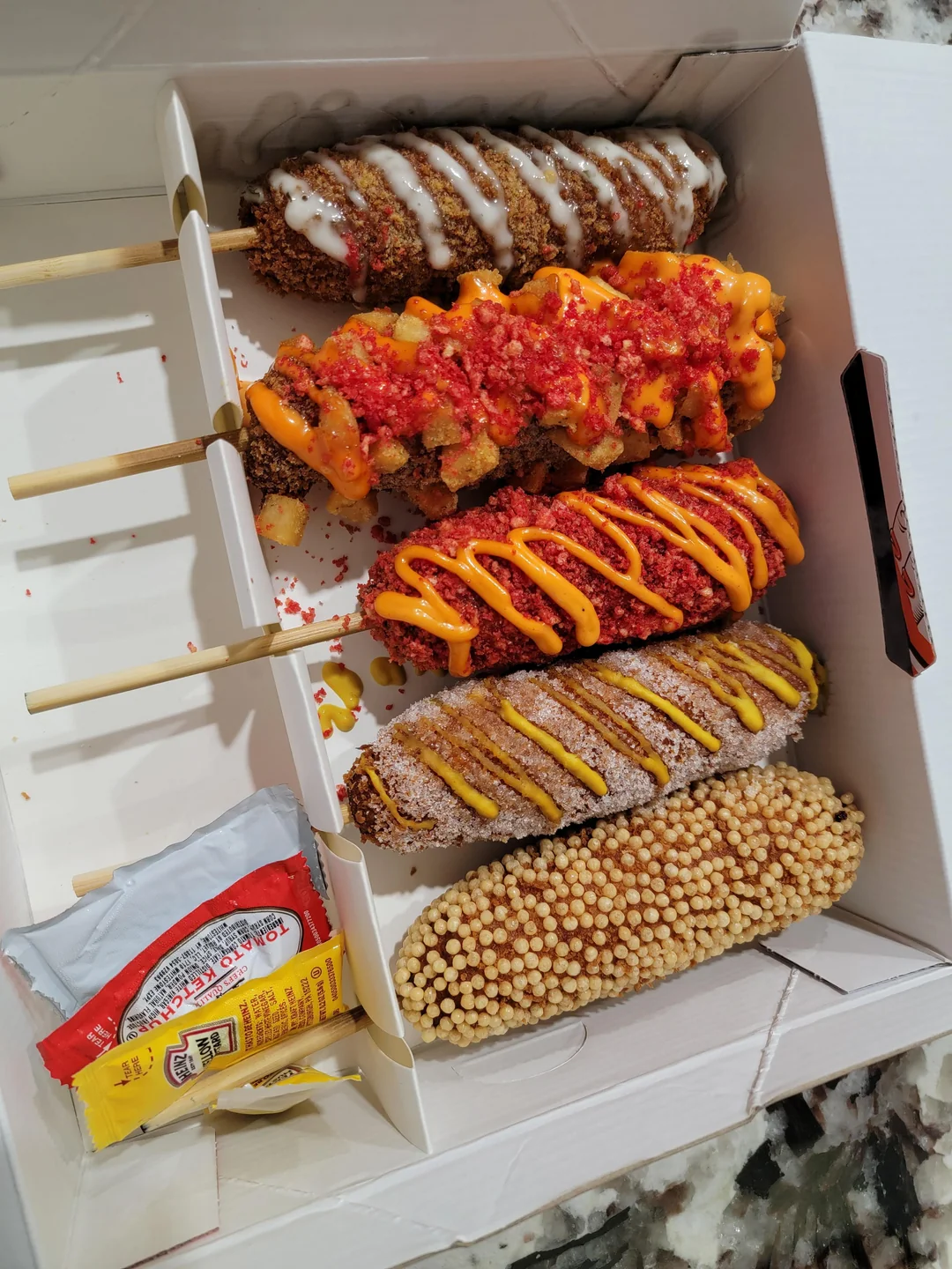 Five Korean corn dogs with different coatings and toppings, including sauces and crumbs, are arranged in a white takeout box. There are also ketchup and mustard packets in the corner of the box.