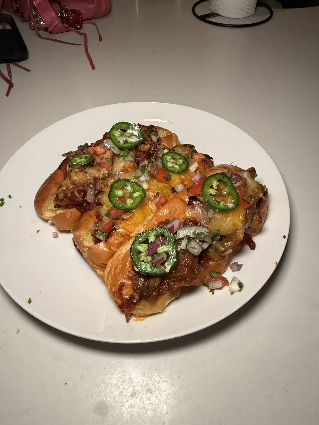 Cheesy loaded bread topped with sliced jalapeños, diced red onion, tomatoes, and herbs is served on a white plate on a light-colored table.