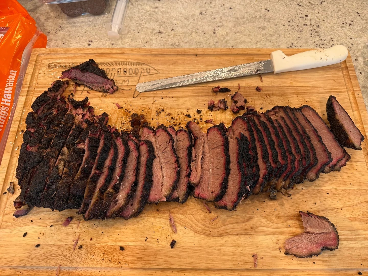 Sliced smoked brisket on a wooden cutting board with a serrated knife and crumbs visible. The brisket has a dark bark and pink smoke ring.