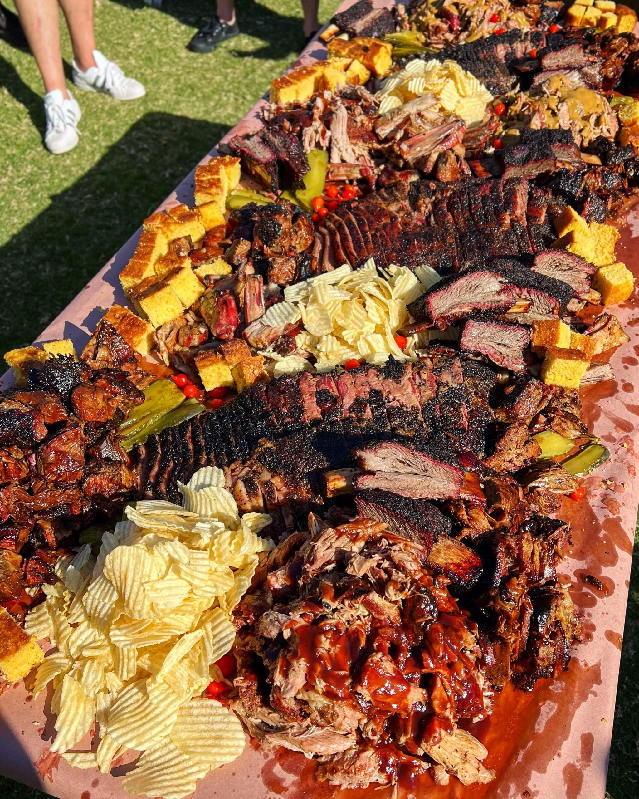 A table covered with barbecue meats, cornbread, pickles, potato chips, and barbecue sauce—everything you need for one of the best BBQ cookouts this summer. Several people stand nearby on grass, visible only from the knees down.