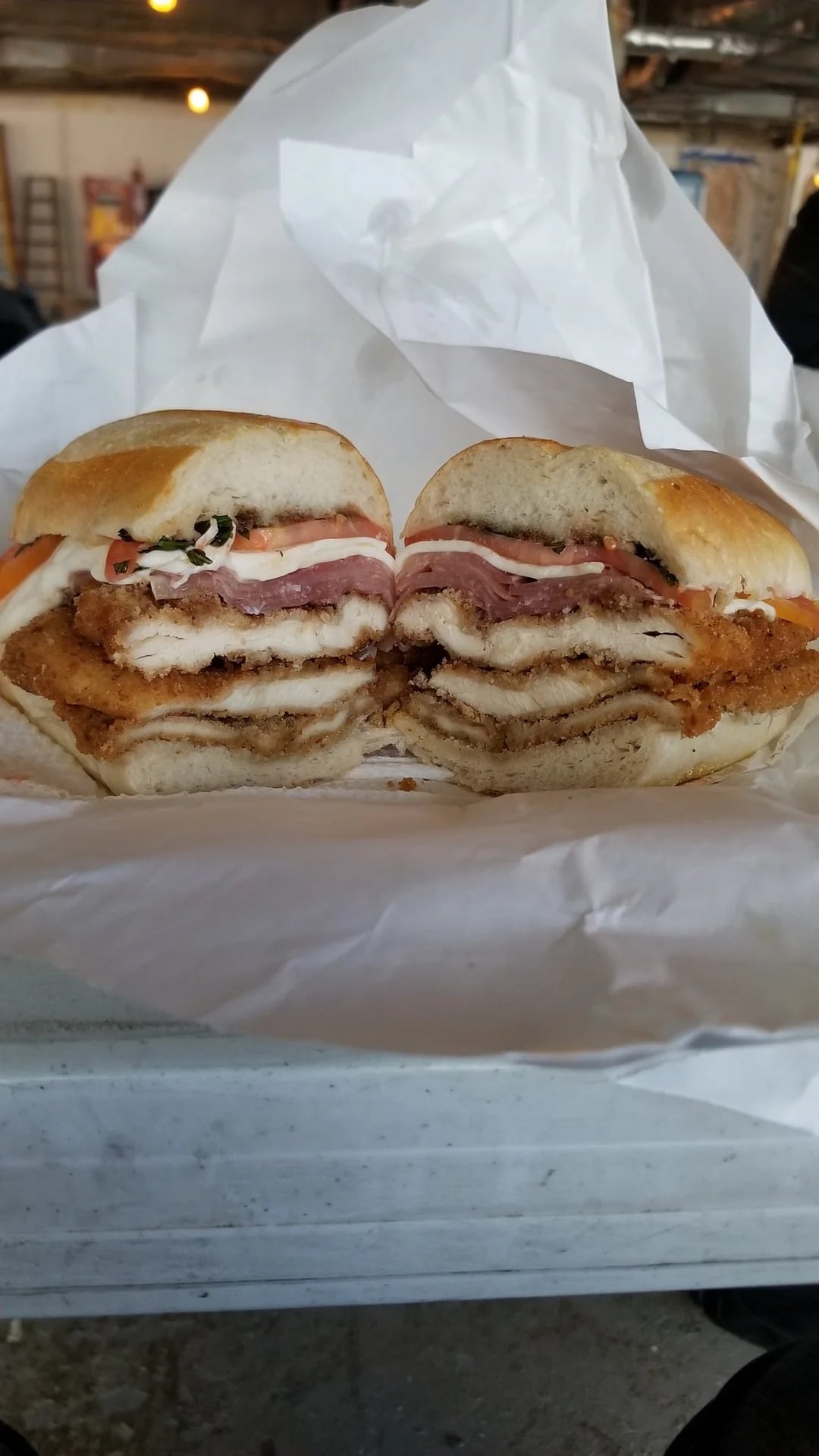 A sandwich cut in half, showing layers of breaded chicken, ham, cheese, tomato, and lettuce, all stacked between a round sandwich bun, wrapped partially in white parchment paper.