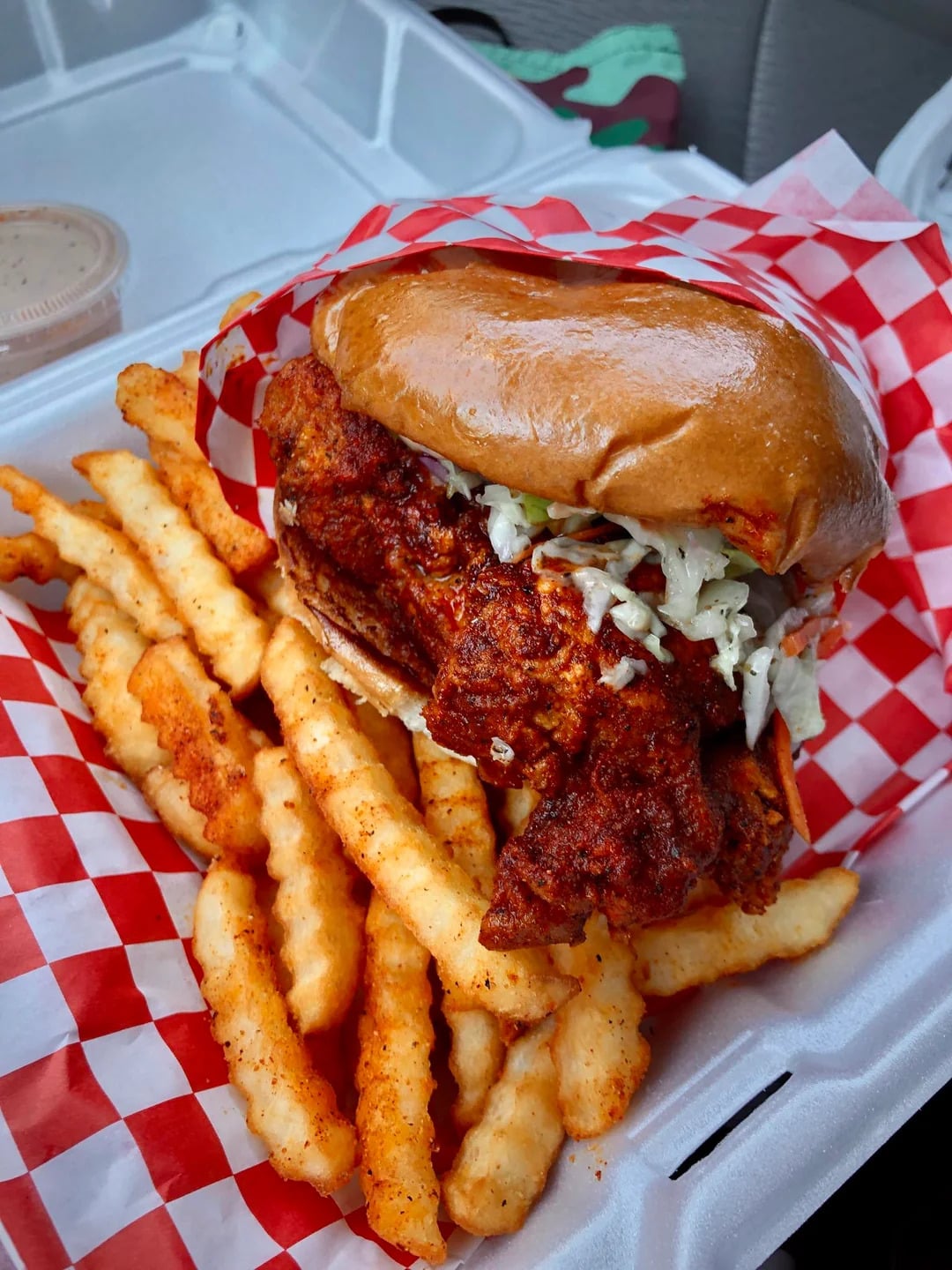 A takeout box with crinkle-cut fries and a fried chicken sandwich topped with coleslaw on a red and white checkered paper. A container of dipping sauce is in the upper corner.