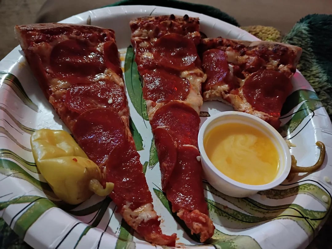 Three slices of pepperoni pizza on a paper plate with a green leaf pattern, a yellow pepper, and a small container of yellow dipping sauce.