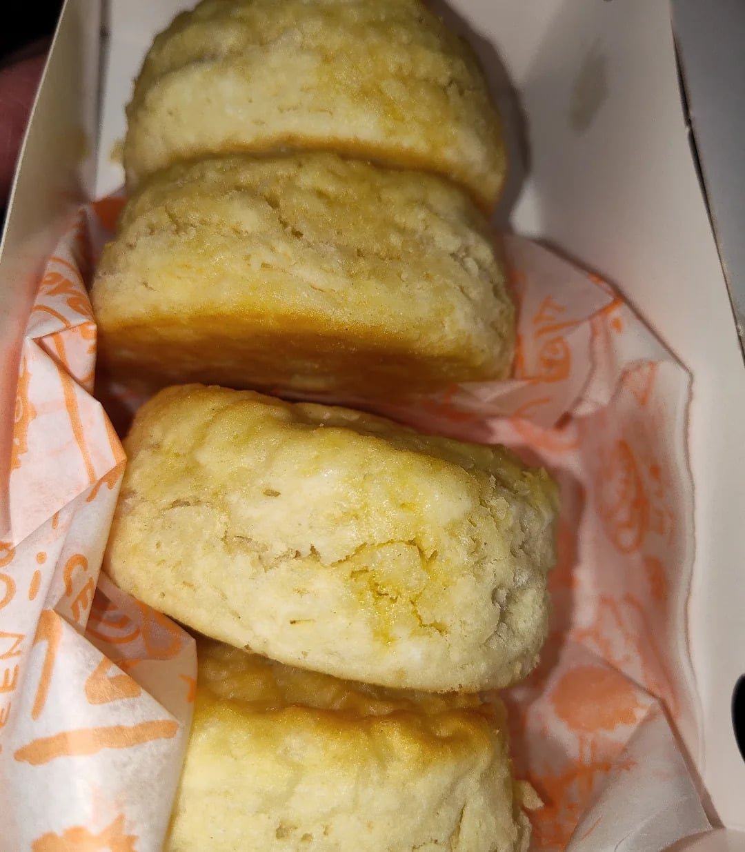 Three golden-brown biscuits are stacked in a takeout box lined with branded orange and white paper. Reminiscent of fast-food favorites, these biscuits look fluffy and slightly crumbly—perfect for enjoying at home or as part of your favorite grocery store products.