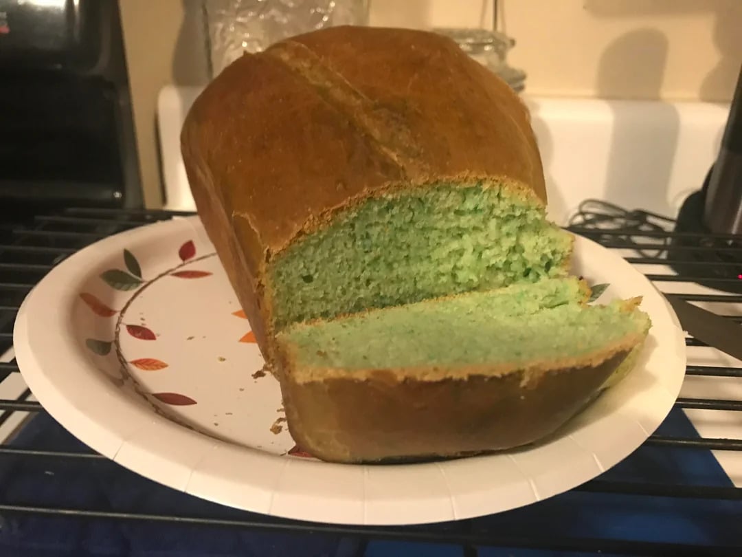 A loaf of bread with a green interior sits on a paper plate decorated with leaves. One slice is cut and slightly separated from the loaf. The bread's crust is golden brown.