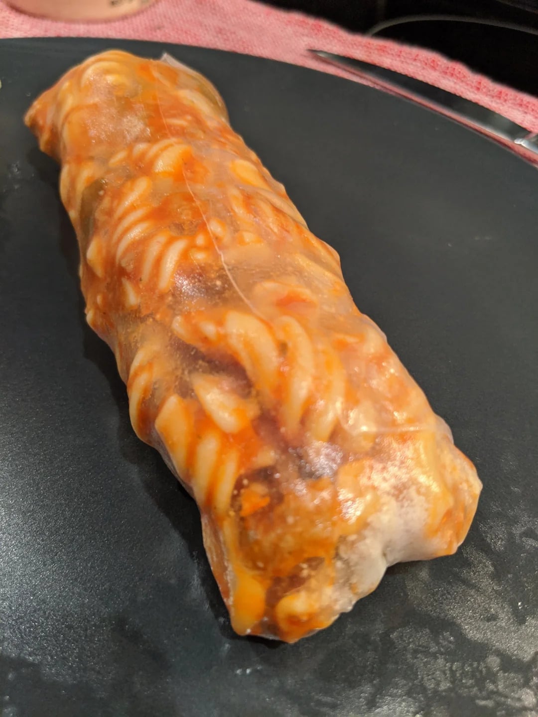 A rice paper roll filled with spiral pasta and red sauce sits on a dark plate.