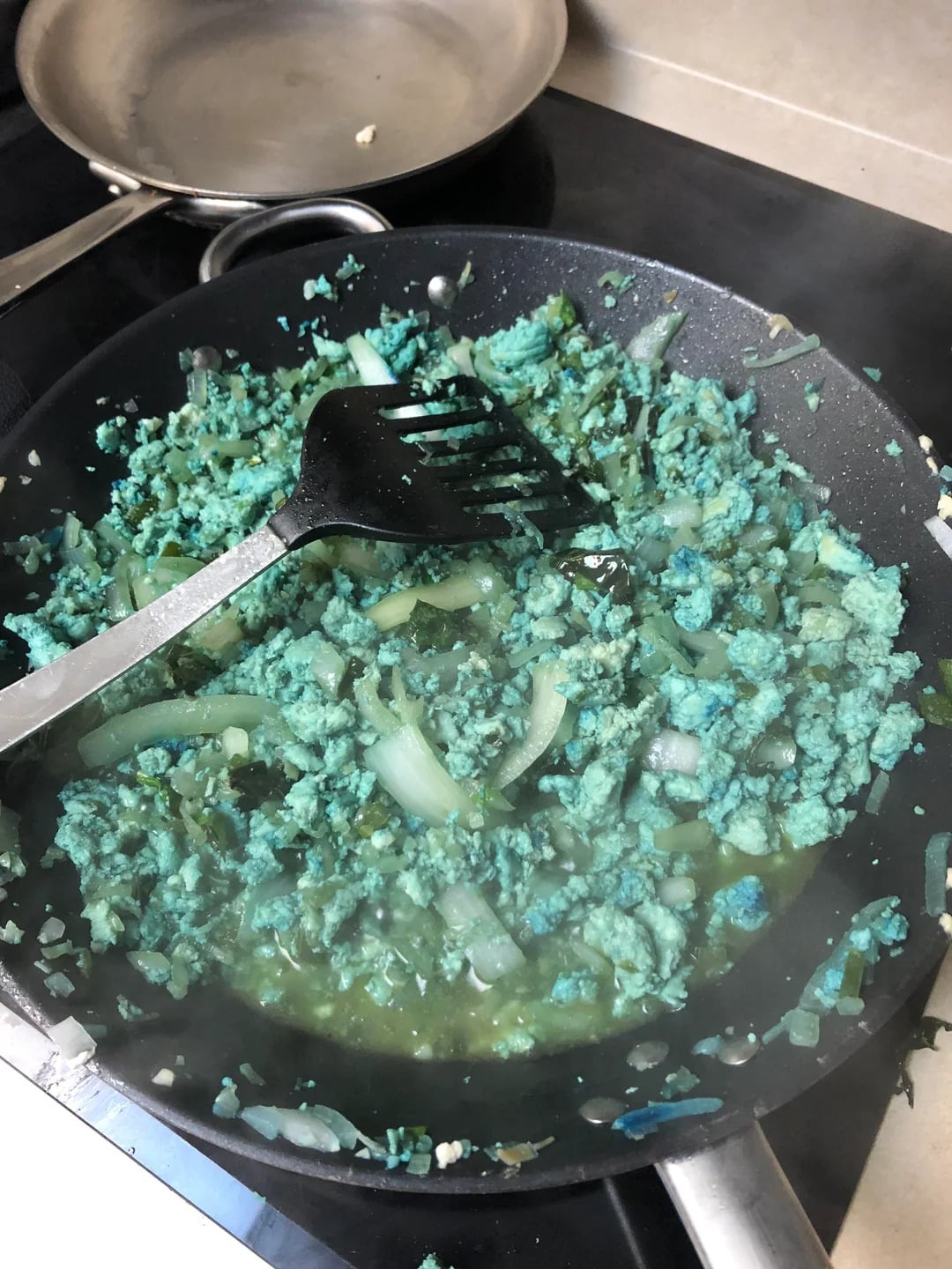 Ground meat and chopped onions are cooking in a black frying pan. The meat has an unusual bright blue-green color. A slotted spatula is resting in the pan, and a second pan is visible in the background.