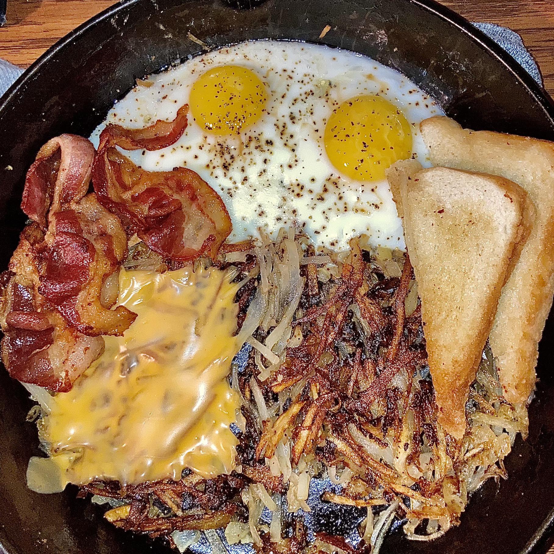 A skillet breakfast with two sunny side up eggs, crispy bacon, hash browns with melted cheese, and two slices of toasted bread.