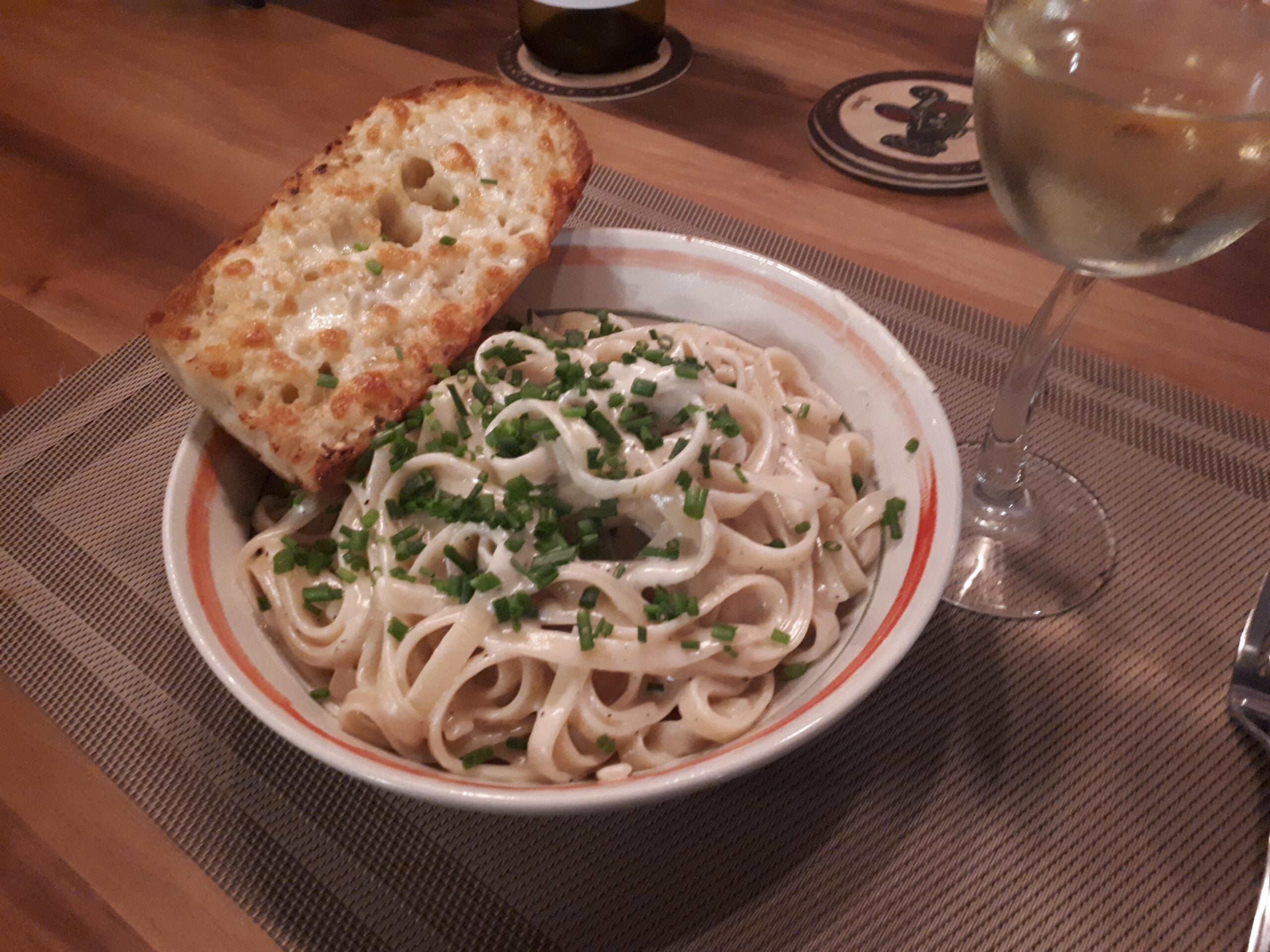 A bowl of fettuccine pasta topped with chopped chives, served with a slice of cheesy garlic bread and a glass of white wine on a wooden table.