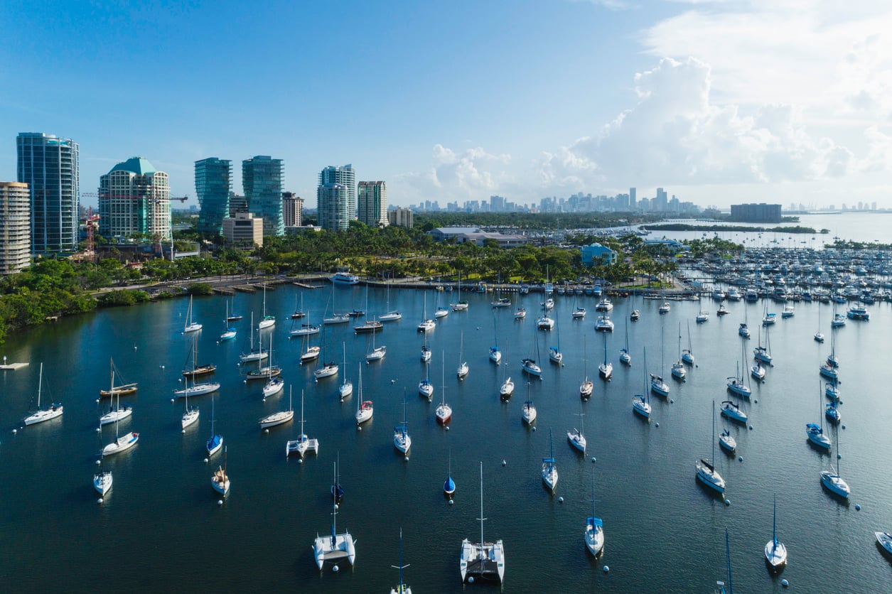 Aerial view of a marina filled with sailboats on calm water, modern high-rise buildings and green spaces along the shoreline, and a city skyline visible in the background under a blue sky with scattered clouds.