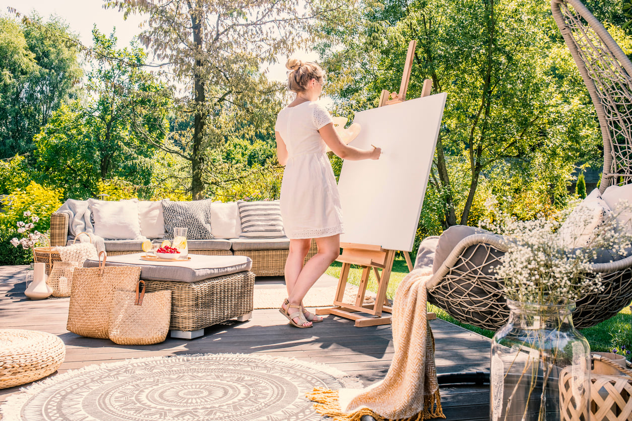 A woman in a white dress stands on a sunny patio, painting on a blank canvas at an easel. Cozy outdoor home products—such as inviting furniture, a rug, and lush greenery—surround her.