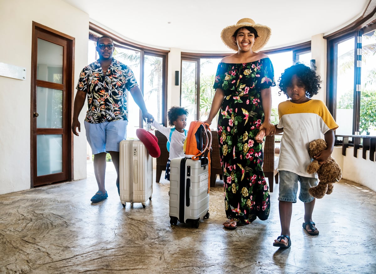 A family of four with suitcases walks into a bright hotel lobby. The parents smile, and the children carry a teddy bear and pillow, suggesting they are on vacation or perhaps enjoying one of their exciting vacation swaps. Large windows show a sunny view outside.