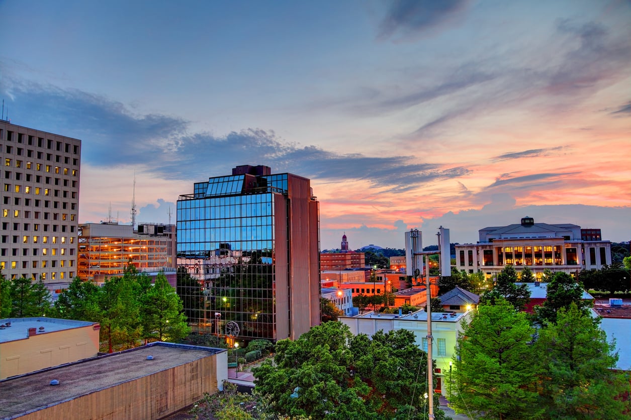 Downtown cityscape at sunset with modern glass buildings, trees in the foreground, and colorful clouds in the sky. A large historic building is visible on the right.