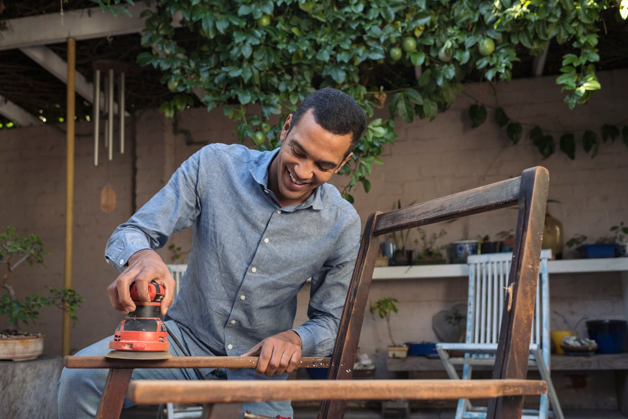 A man smiles while sanding a wooden chair with an electric sander outdoors, surrounded by plants and garden furniture.