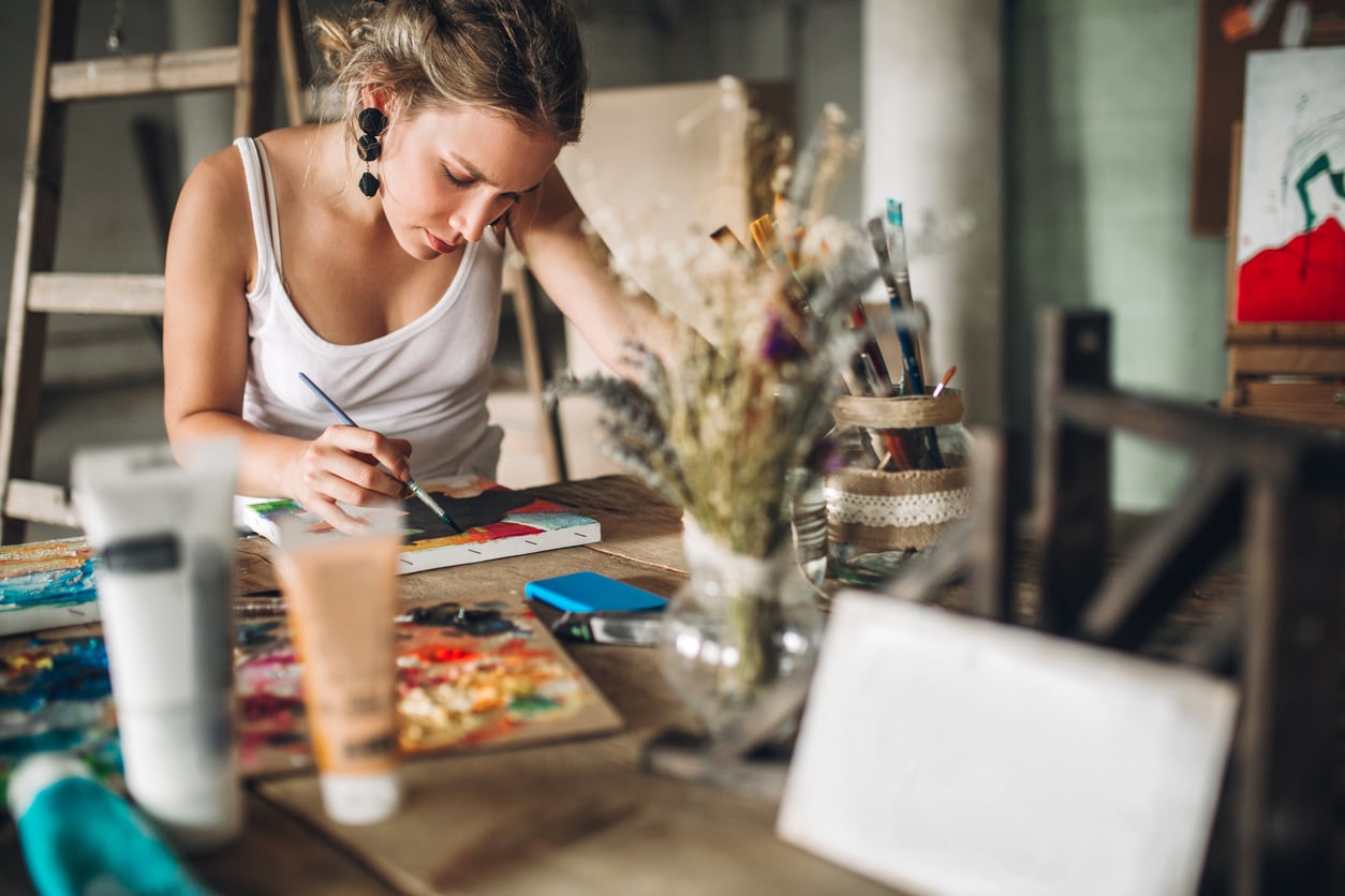 A woman in a white tank top sits at a wooden table painting on a canvas, surrounded by art supplies, brushes, tubes of paint, and a vase with dried flowers—a quiet scene that evokes life before smartphones, when creativity flourished uninterrupted.