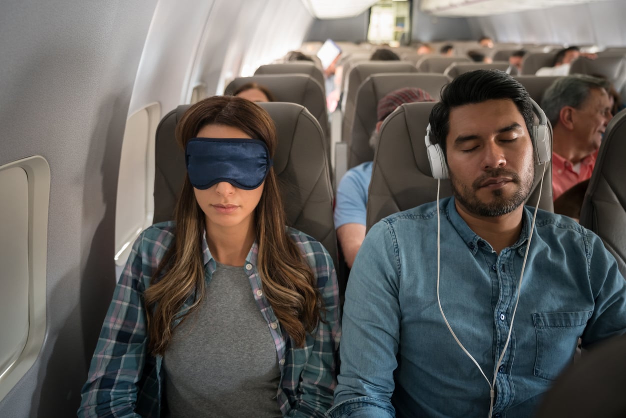 A woman wearing a sleep mask and a man with headphones sit side by side, both resting, on an airplane with other passengers seated behind them.