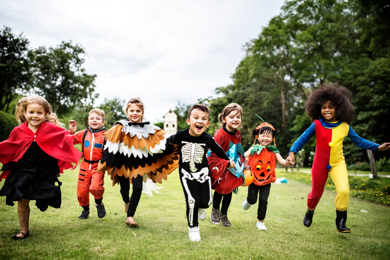 Seven children in colorful costumes, including a skeleton, pumpkin, and bird, run and smile together on a grassy lawn outdoors, enjoying Halloween activities with trees and a bright sky in the background.