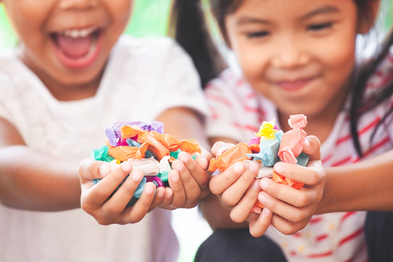 Two children smile and hold out colorful wrapped candies in their hands, with faces slightly blurred and the focus on the candy.