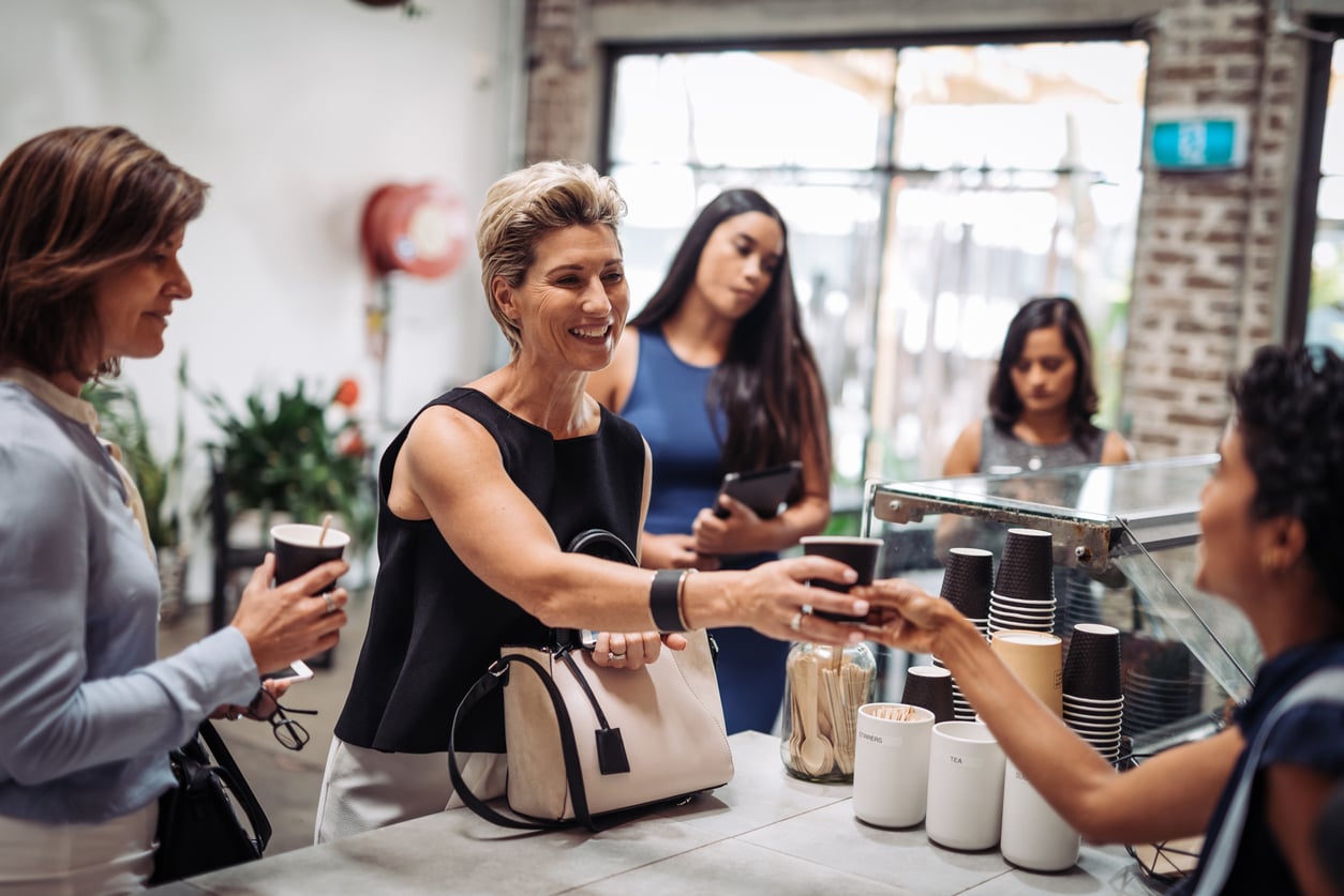A smiling woman receives a coffee cup from a barista at a café counter, while others wait in line behind her, holding drinks and chatting. The setting is bright and modern—a scene that reflects how women are changing their spending behaviors in today’s vibrant social spaces.