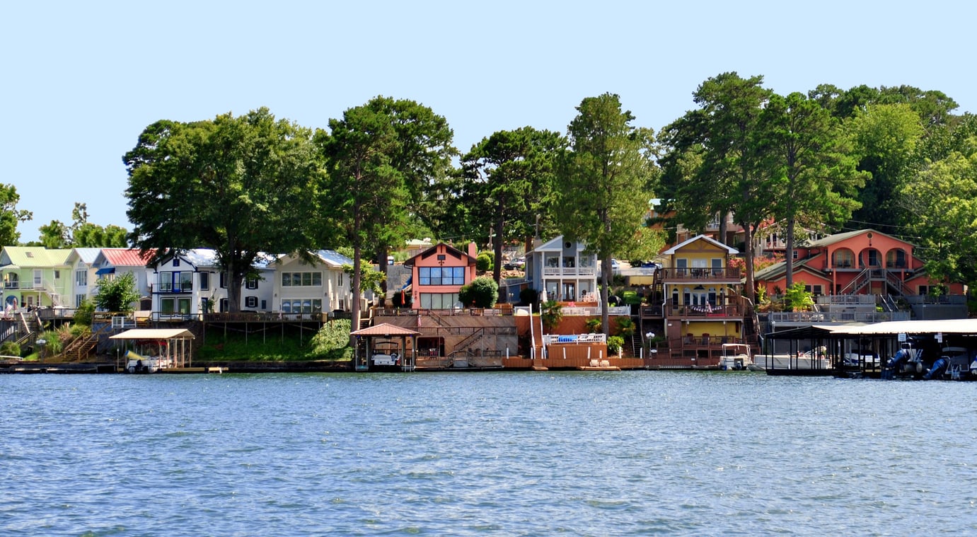Colorful lakeside houses surrounded by trees, with boat docks and calm blue water in the foreground under a clear sky.