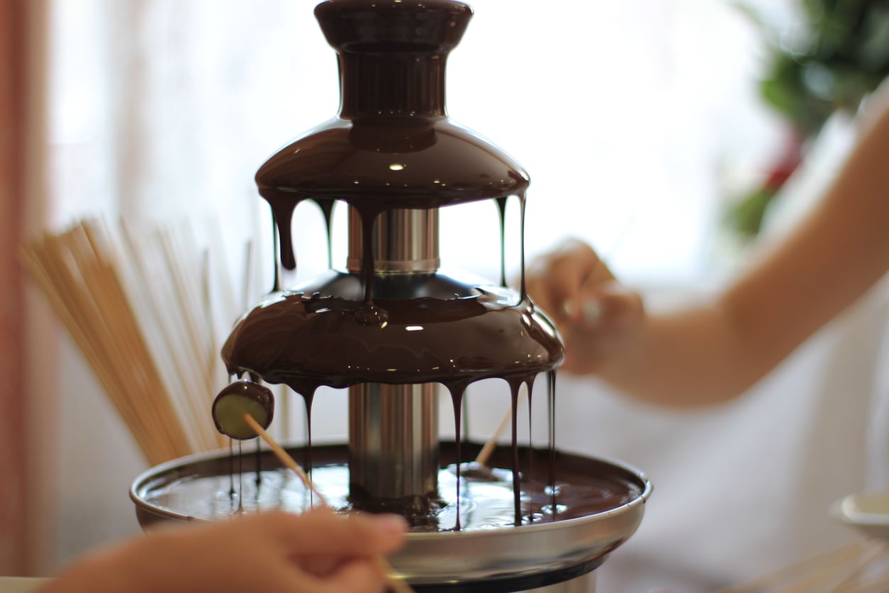 A chocolate fountain with flowing chocolate, surrounded by people dipping skewers, likely for a dessert or fondue experience, showcases one of the most delightful kitchen gadgets in action. Blurred background with skewers visible.