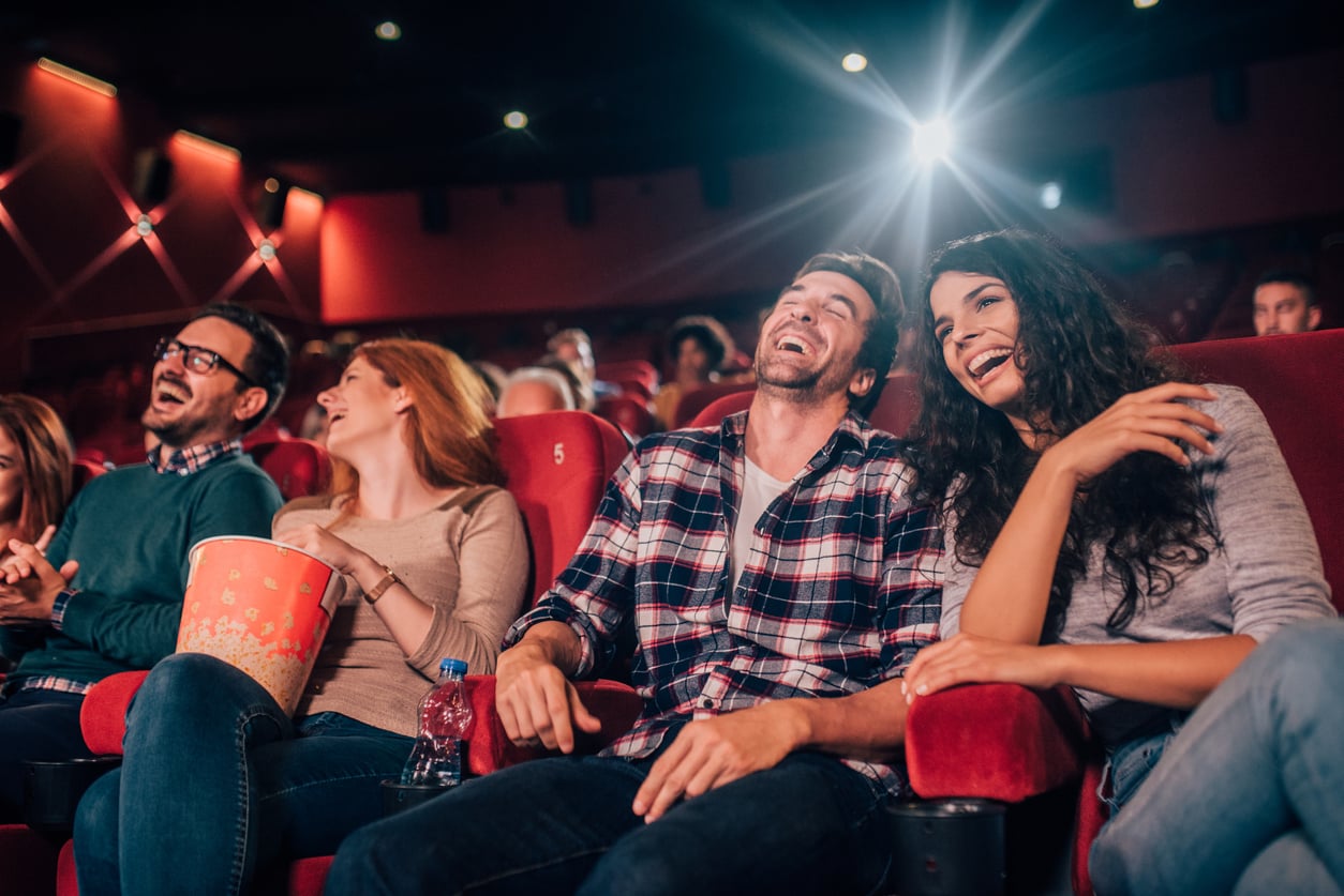 Four people sit in red theater seats, laughing and enjoying a movie. One person holds a large popcorn bucket, ready to share some movie theater hacks for the best experience. The dim theater glows with bright projector light from behind them.