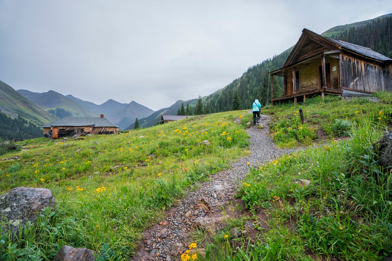 A person walks on a gravel path toward a rustic wooden cabin in a green meadow with wildflowers, reminiscent of the peaceful charm found in many small American towns, surrounded by mountains and cloudy skies.