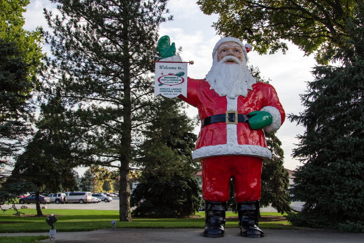 A large statue of Santa Claus stands outdoors, holding a “Welcome” sign—a fitting sight for one of the best Christmas towns in America. Santa is surrounded by pine trees, and cars are parked in the background under a partly cloudy sky.