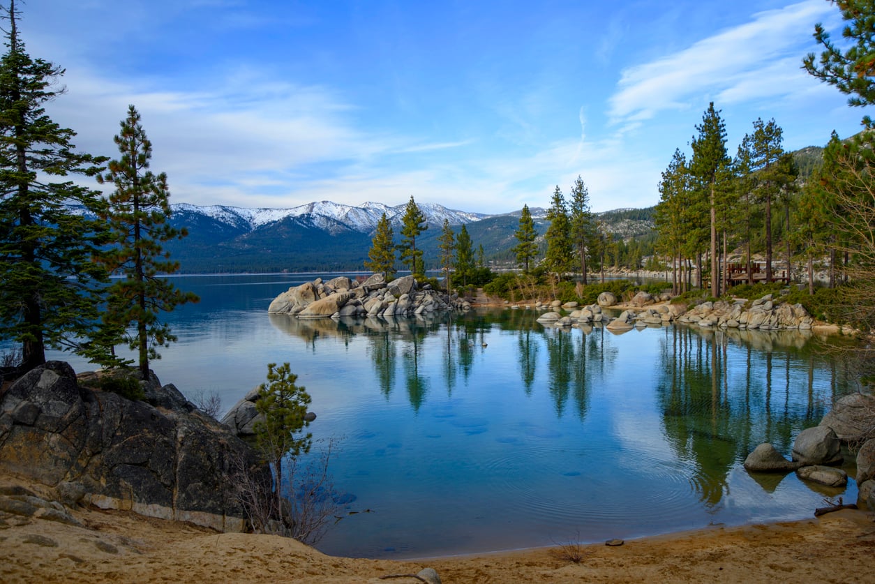 A calm lake surrounded by pine trees and large rocks, with snow-capped mountains in the background under a partly cloudy blue sky.