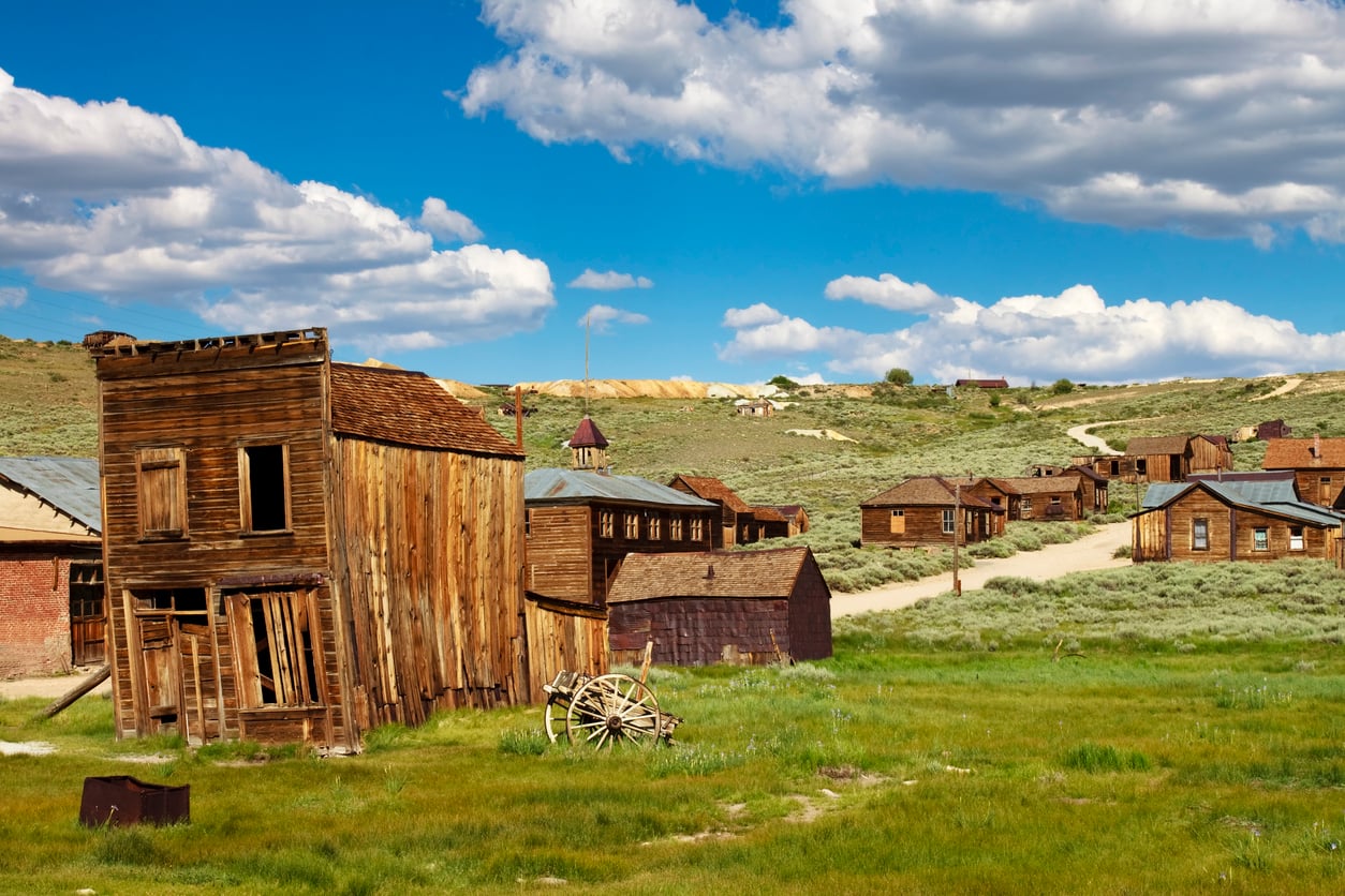 A deserted Old West town with worn wooden buildings, a broken wagon wheel, and grassy, open land under a partly cloudy blue sky captures the haunting spirit of early American towns.