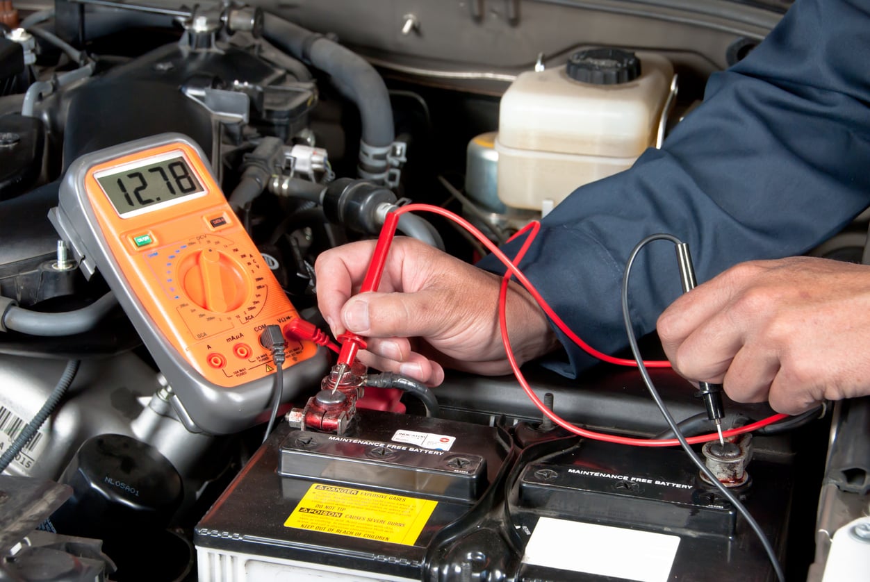 A person uses a digital multimeter to check the voltage of a car battery under the hood, with the meter displaying 12.78 volts—an essential step on any thorough road trip checklist.