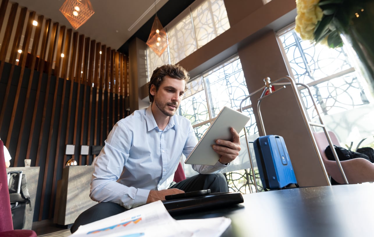 A man in a light blue shirt sits in a modern hotel lobby, looking at a tablet—perhaps reviewing some hotel booking tips before his next trip. A blue suitcase is beside him, and there are papers and a laptop on the table in front of him.