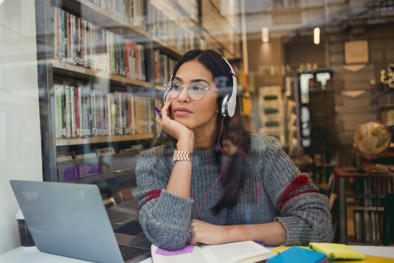 A woman wearing headphones sits at a library table with a laptop and open notebook, looking thoughtfully out the window. Bookshelves are visible in the background.