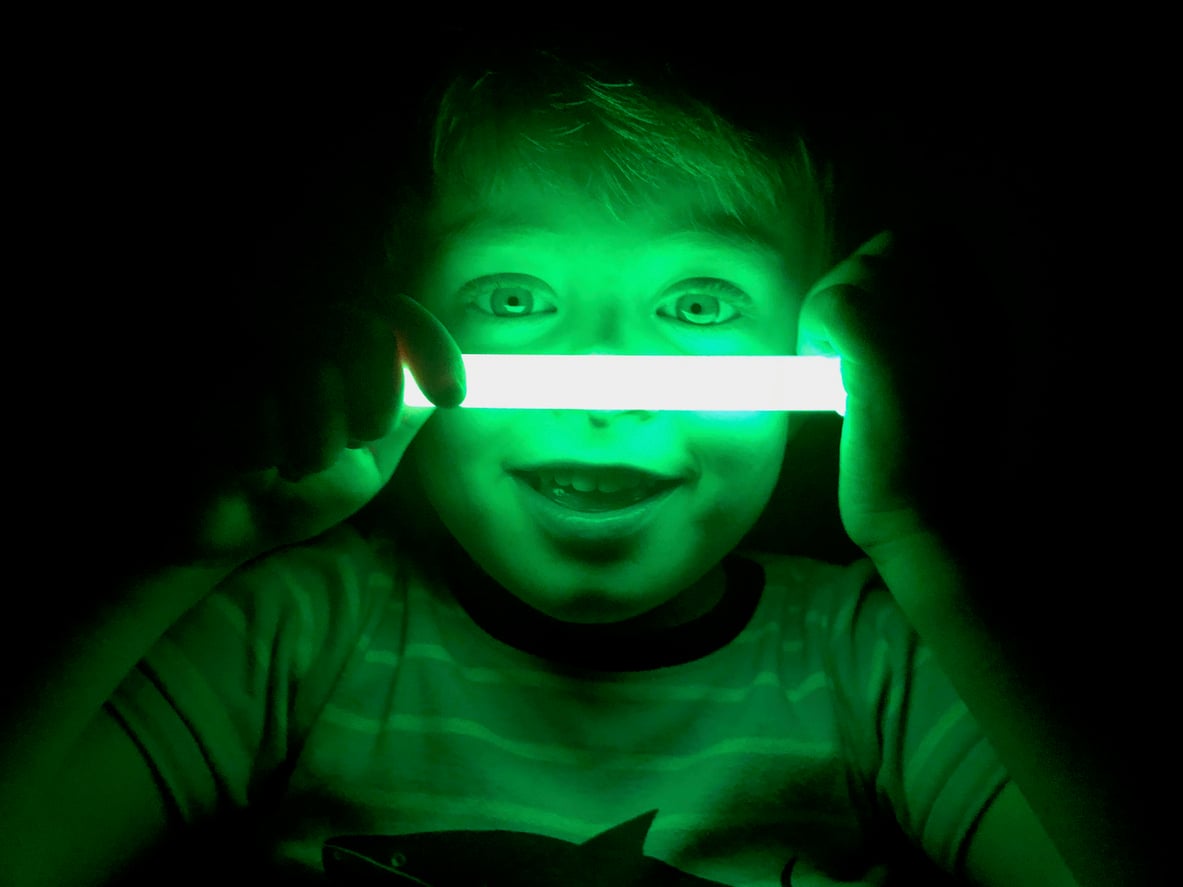A child holding a glowing green stick in a dark room, illuminating their smiling face and eyes—capturing the excitement and wonder of Halloween activities.