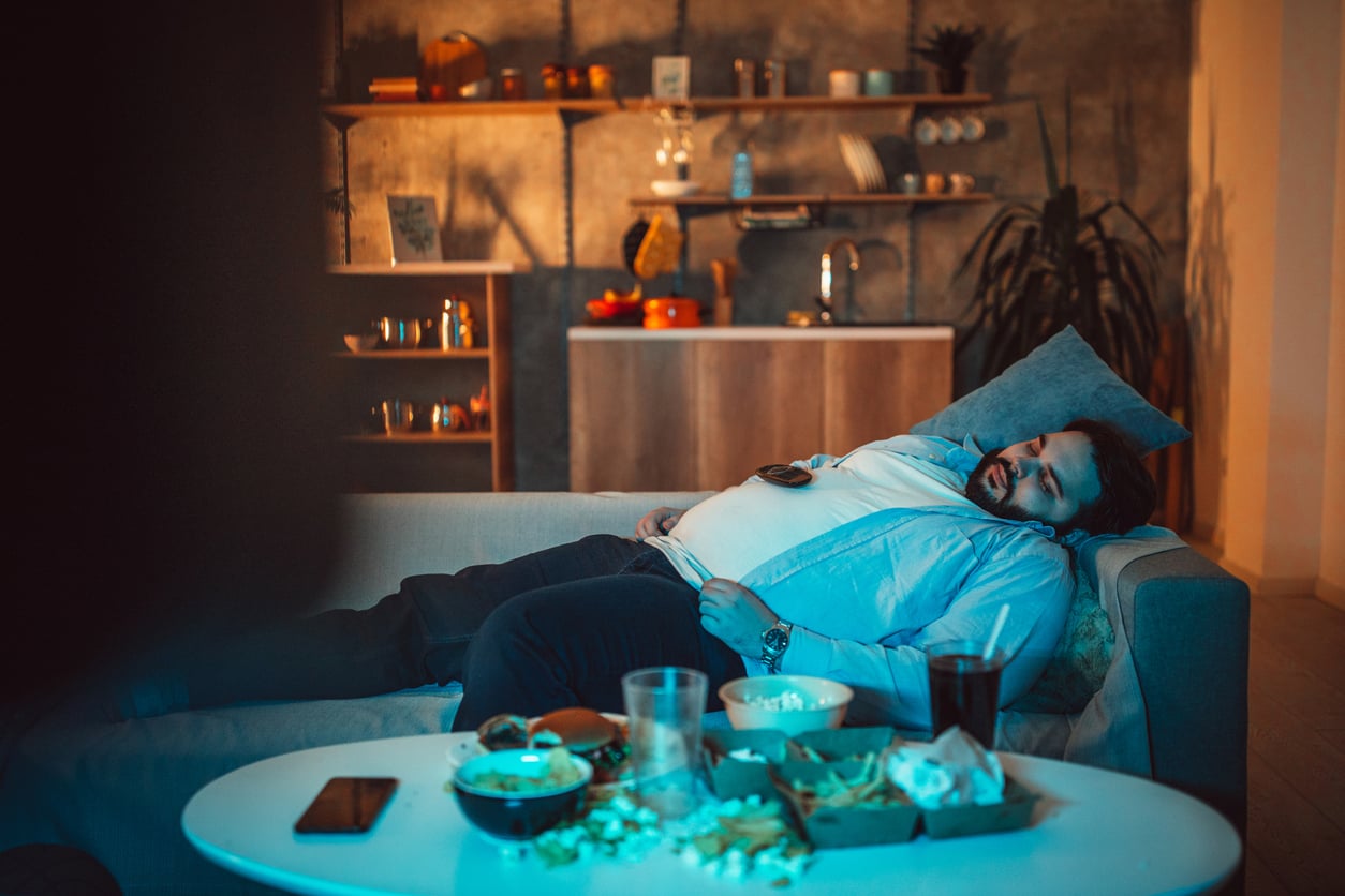 A man is sleeping on a couch in a dimly lit living room surrounded by snacks, popcorn, and a remote, perhaps unwinding after a long day of managing household expenses. The kitchen area can be seen in the background.