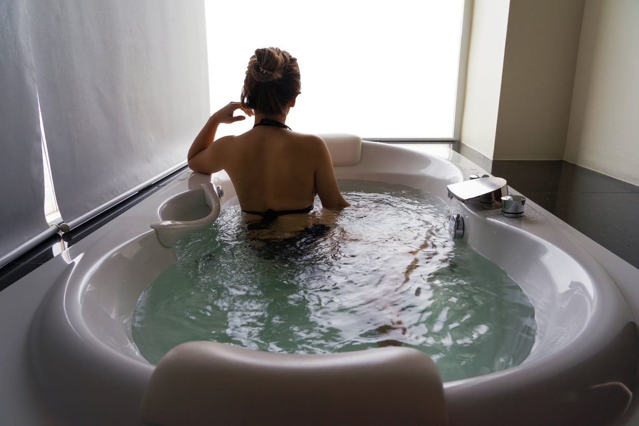 A woman with her hair in a bun relaxes in a round bathtub filled with water, facing a frosted window with natural lightāa serene scene that calls to mind home features homeowners no longer want, as round bathtubs have gradually fallen out of modern design trends. A cup and saucer sit on the tub's edge.