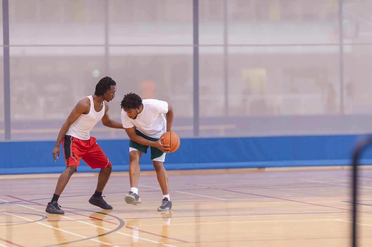 Two young men play basketball indoors; one dribbles the ball while the other defends closely. Both are wearing athletic clothes and sneakers on a polished gym court, making the most of memberships worth paying for.