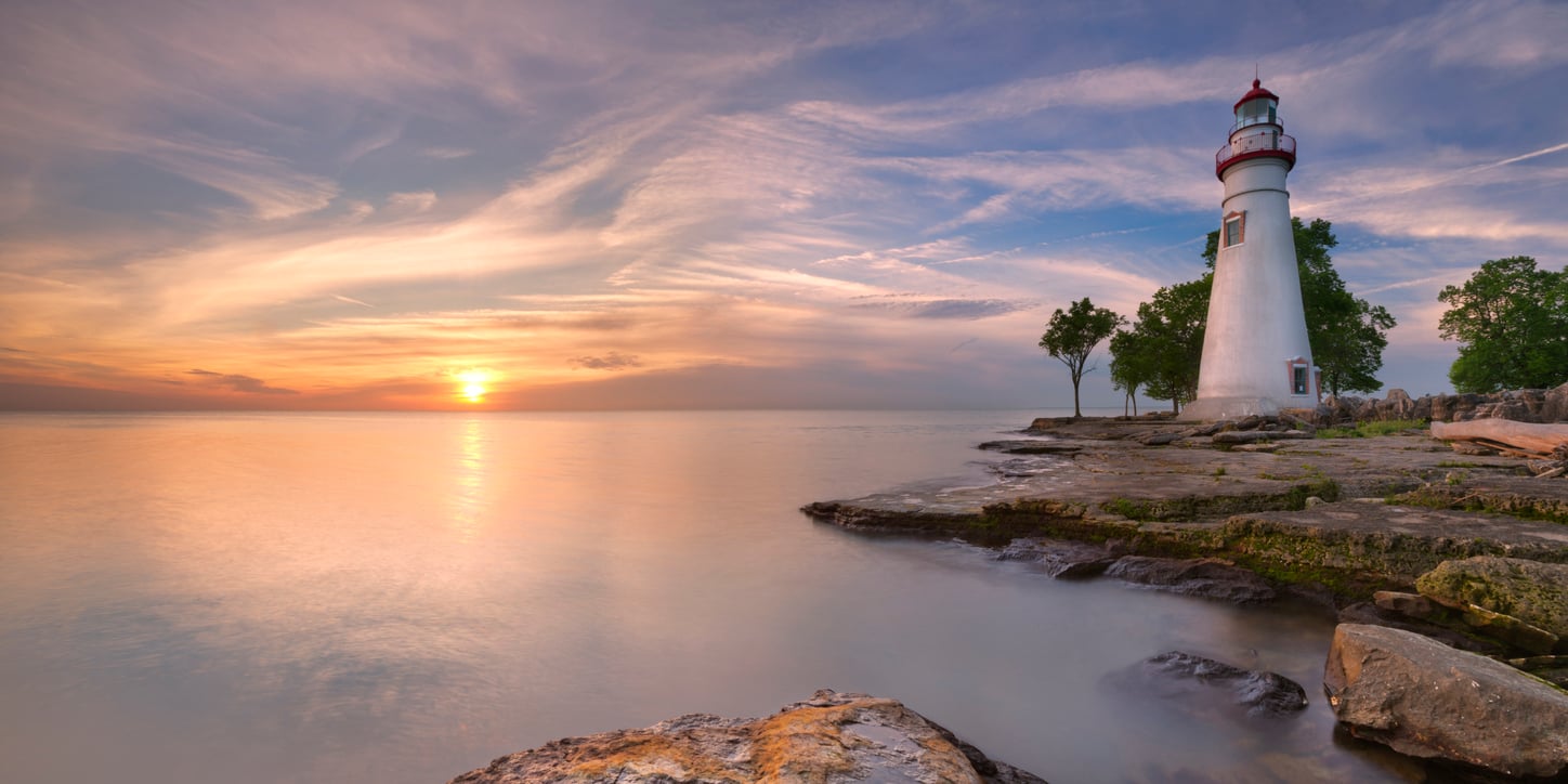 A lighthouse stands on a rocky shoreline with trees nearby, overlooking calm water at sunset under a sky with light clouds.