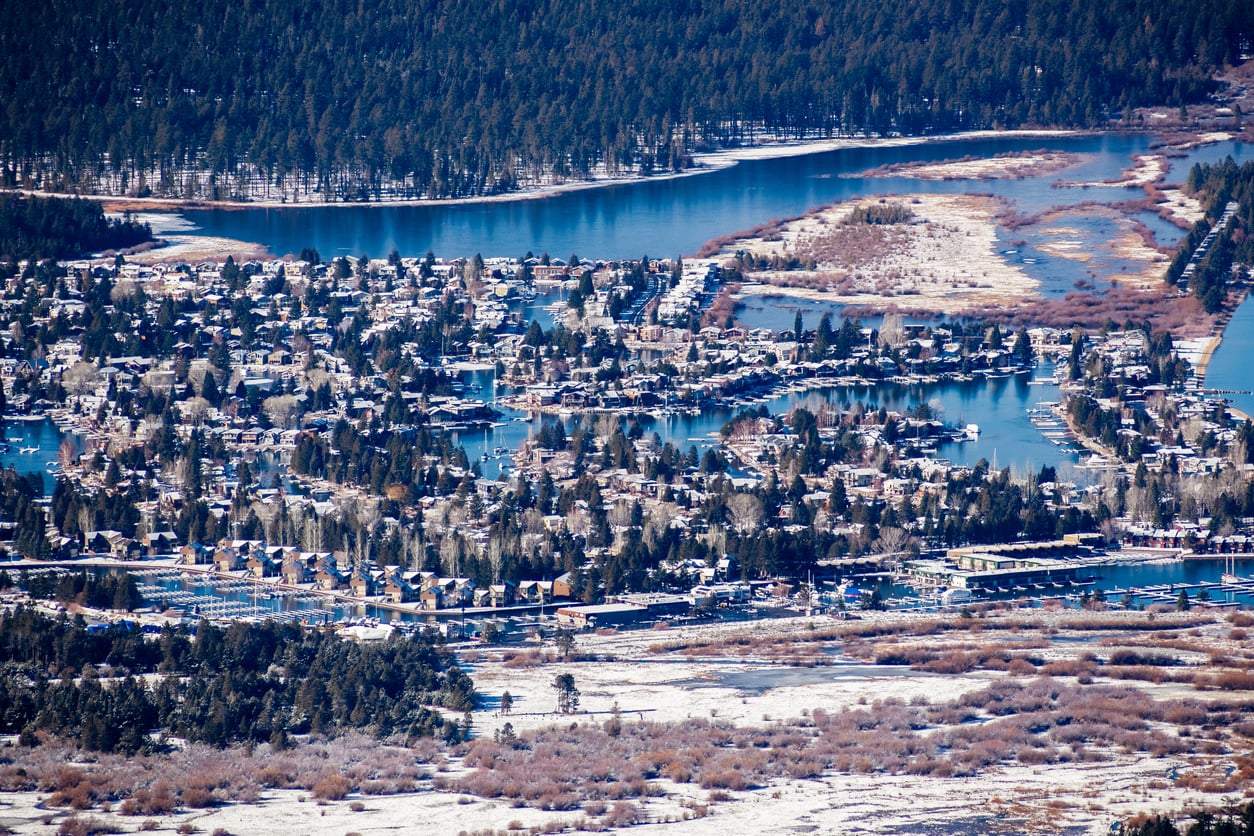 Aerial view of a snowy lakeside community with winding waterways, houses nestled among trees, and forested hills in the background—one of the most picturesque and affordable travel destinations for those seeking a serene winter escape.